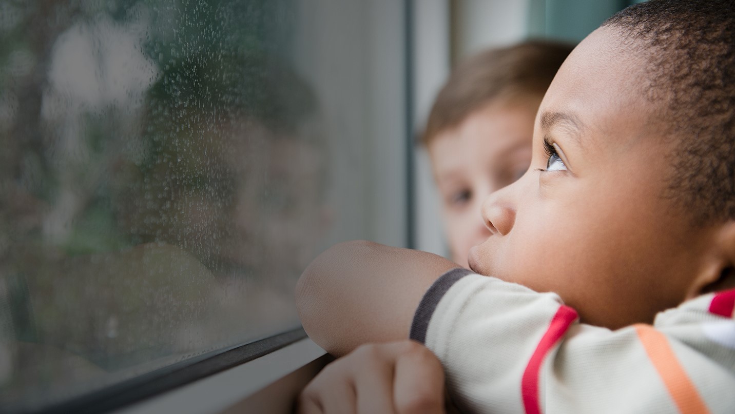 Two young boys looking out a rain drop covered window.
